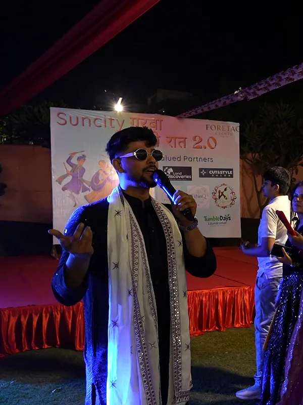 Ashish on the mic beside the partners backdrop as guests line up with dandiya sticks